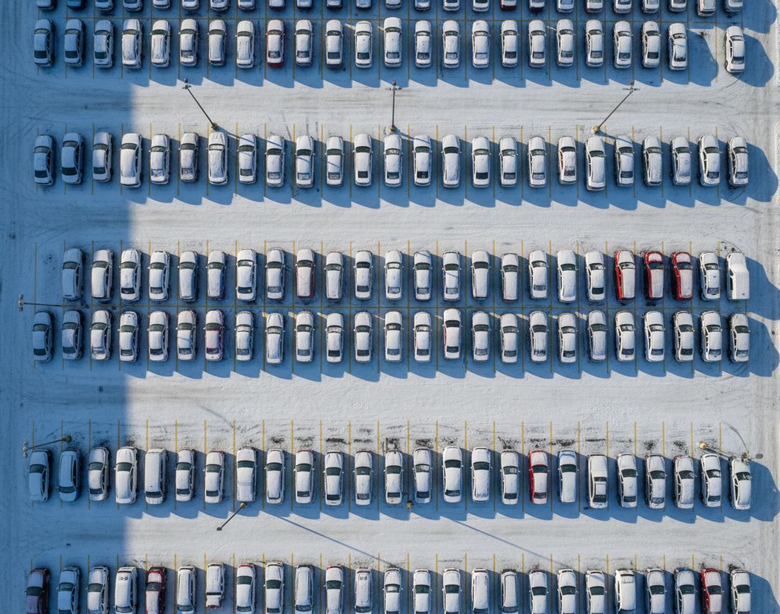 Parked cars seen straight down from above, their roofs dusted with snow, in long rows between yellow lines and the shadows of lamp posts